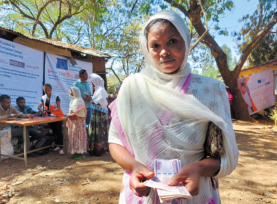 A woman looks at the camera, holding cash. Behind her, people wait in line to receive cash and vouchers.