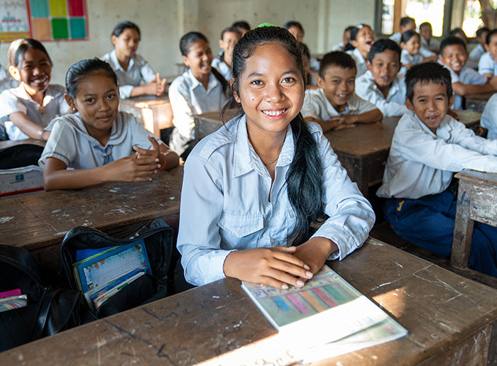 A classroom of smiling students
in uniforms sit at wooden desks, with one girl prominently in the foreground holding a book.