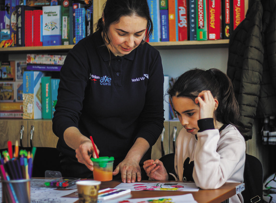 A woman distributes art supplies to a girl sitting at a table with her hand resting against her head.