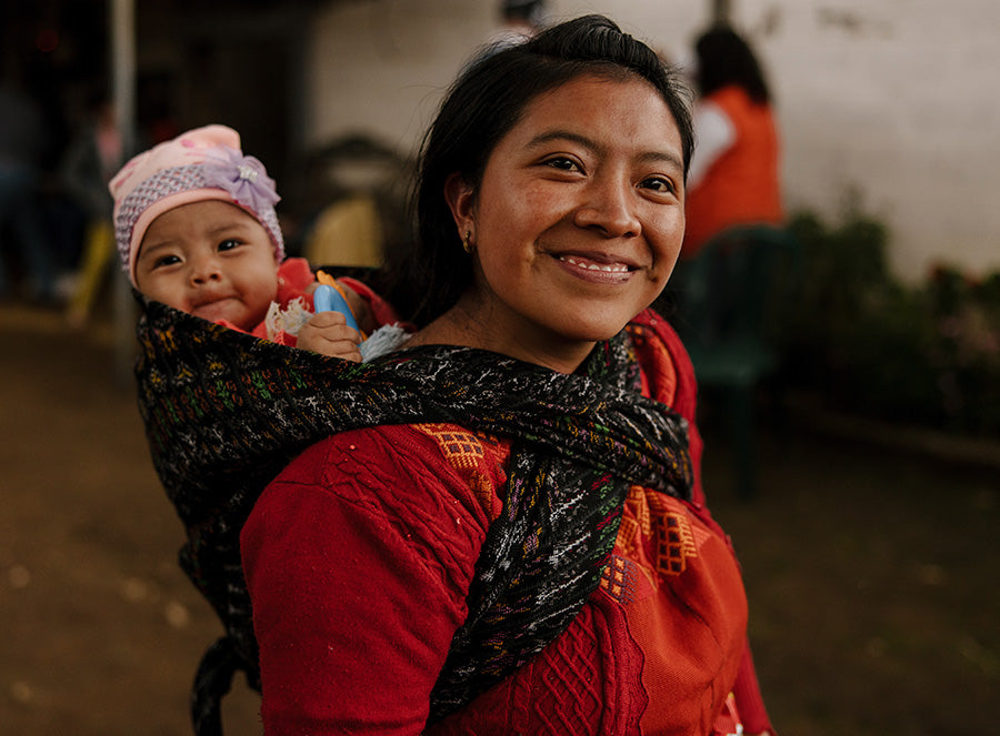 A woman wearing a red shirt with a baby wrapped around her back smiles as the baby peeks over her shoulder.