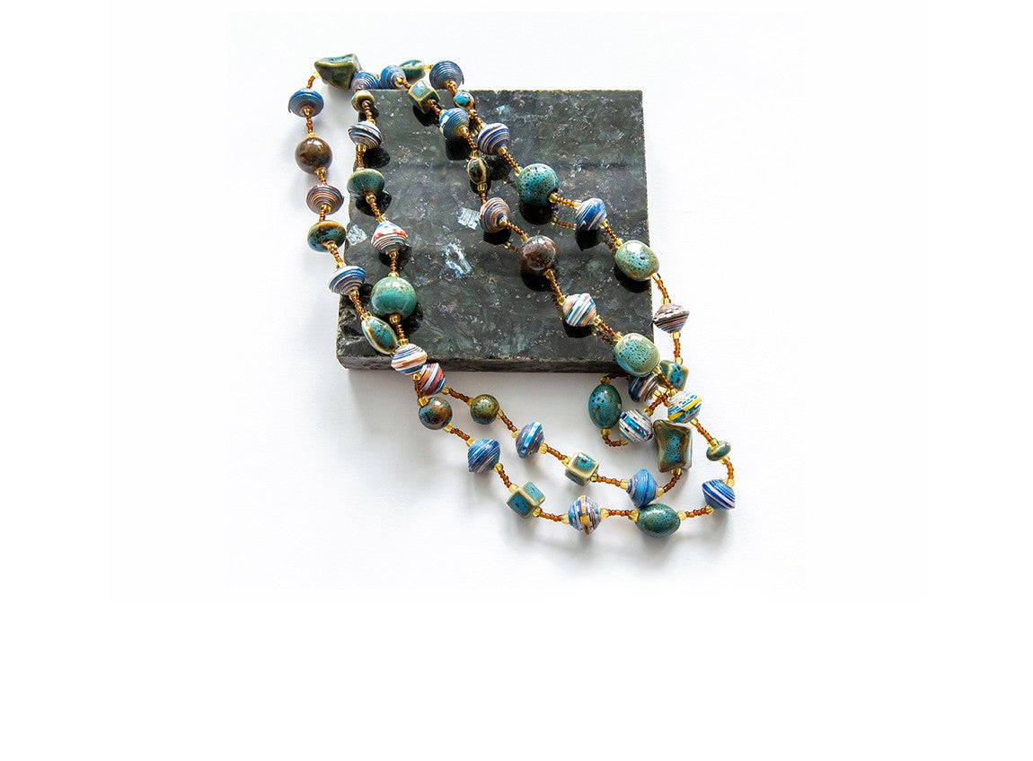 Handcrafted beaded necklace with a mix of round and disc-shaped beads in earthy tones of blue, green, brown, and white, displayed on a dark stone tile against a white background.