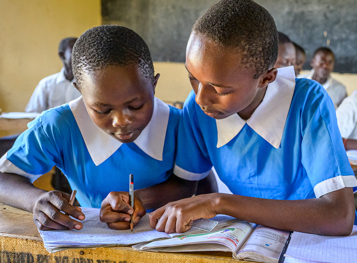 Two girls in blue uniforms sit at a classroom desk, helping eachother with work.