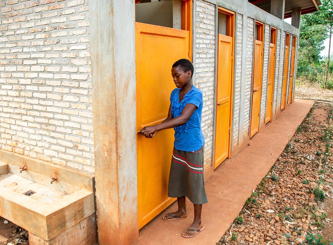 A girl wearing a skirt and blue top closes the door to a stall in a bathrom facility.