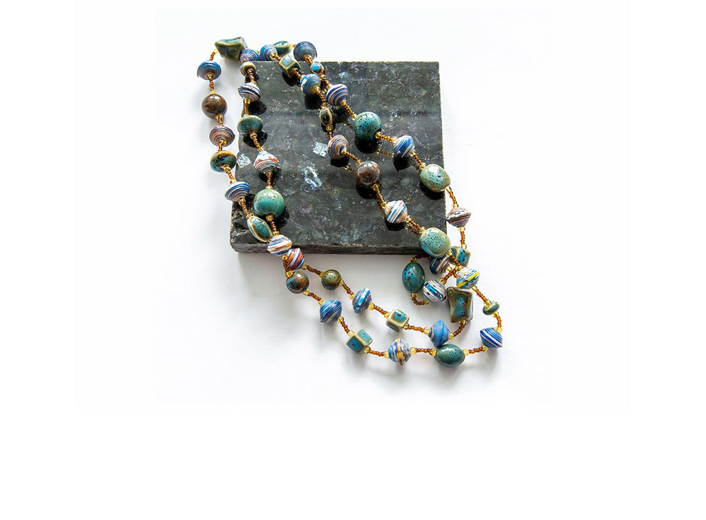 Handcrafted beaded necklace with a mix of round and disc-shaped beads in earthy tones of blue, green, brown, and white, displayed on a dark stone tile against a white background.